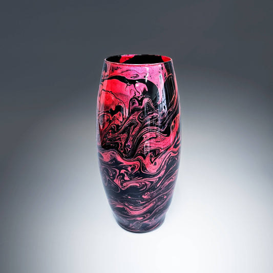This hand-painted, fluid art, modern glass vase is an electric display of vibrant colors swirling together in an elegant dance of metallic rose red on a base of glossy black. The fluidity of the design creates a captivating and unique pattern,