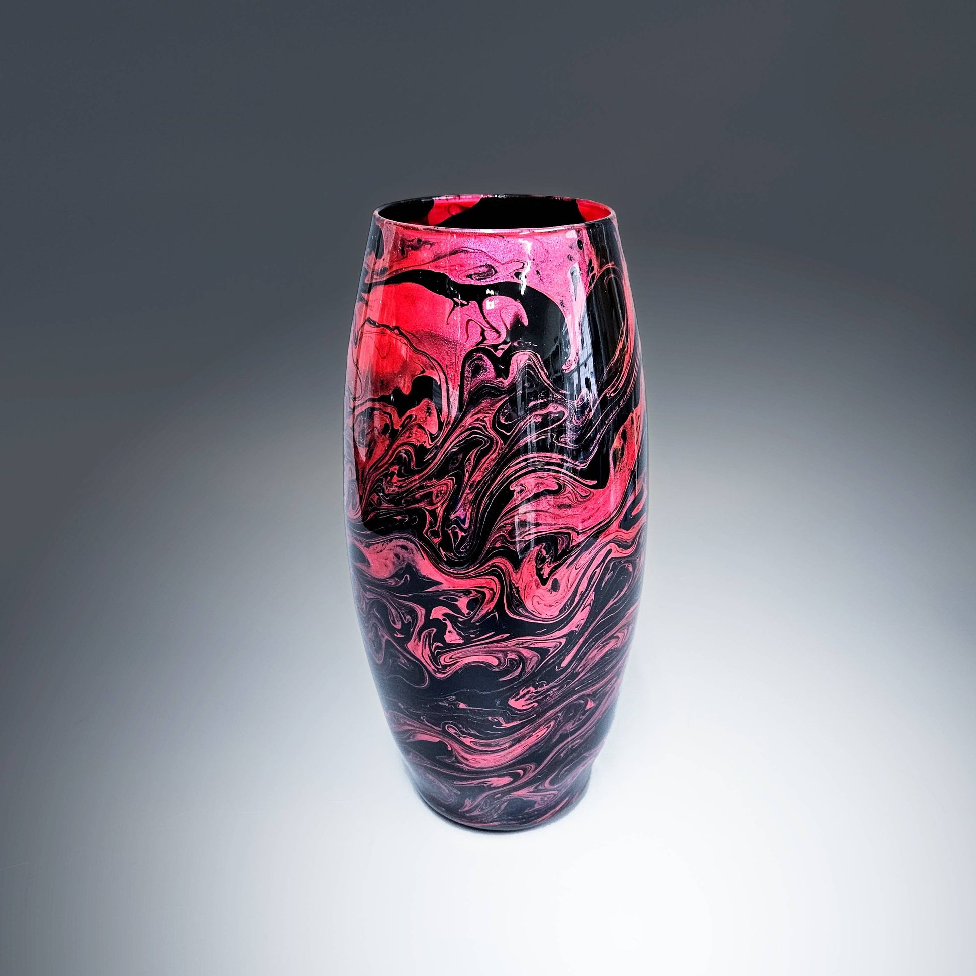 This hand-painted, fluid art, modern glass vase is an electric display of vibrant colors swirling together in an elegant dance of metallic rose red on a base of glossy black. The fluidity of the design creates a captivating and unique pattern,